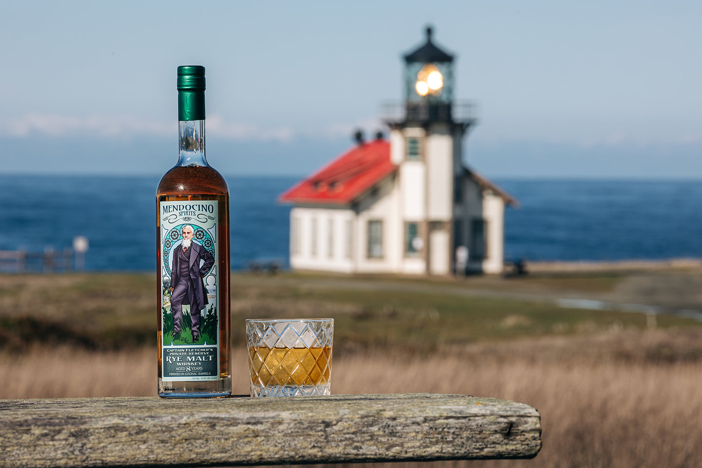 Bottle of Mendocino Spirits Rye Whiskey with a glass on a wooden surface in front of a lighthouse.